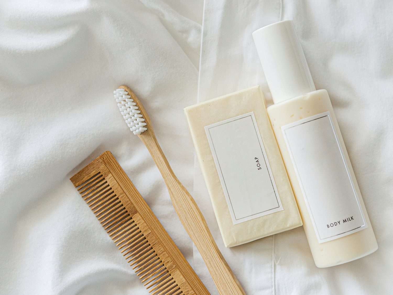 Wooden comb, toothbrush, wrapped soap and body milk laid out on white hotel linen