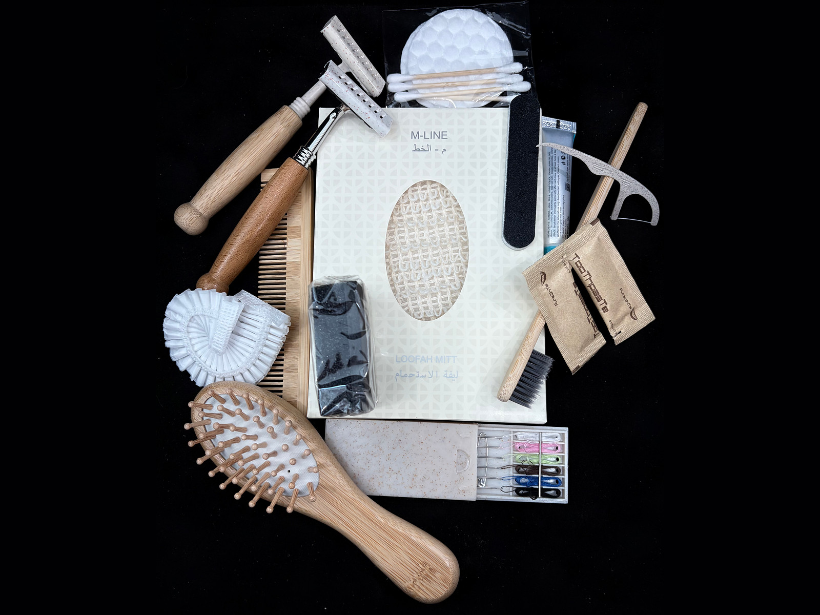 The M-Line dry amenity range laid out around a boxed loofah mitt: bamboo comb and hairbrush, razors, shower cap, cosmetic pads and cotton buds, nail file, bamboo toothbrush, toothpaste, mending kit and shoeshine sponge