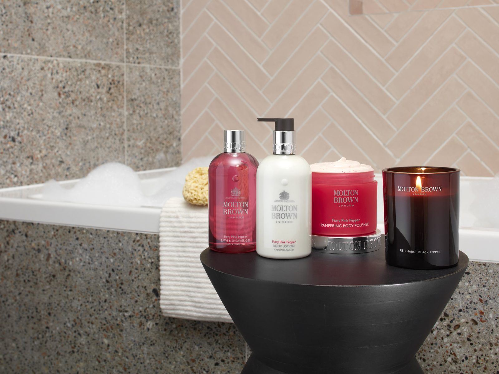 Molton Brown bath and body products and a candle on a side table beside a bath