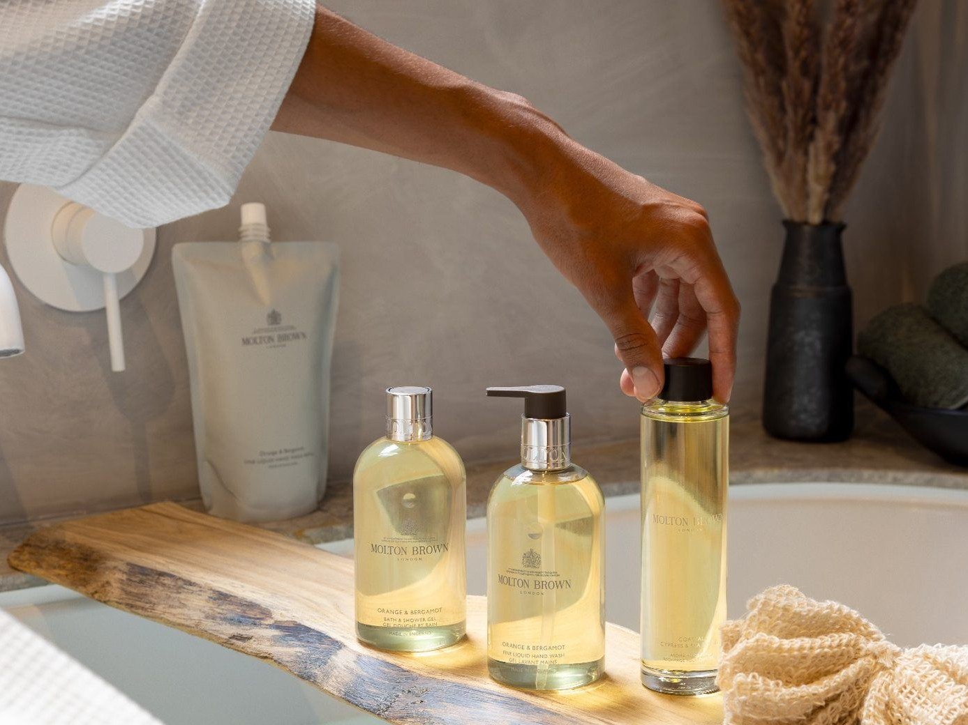 A guest reaching for a Molton Brown bath oil on a timber caddy across a bath, with the 5L refill pouch behind
